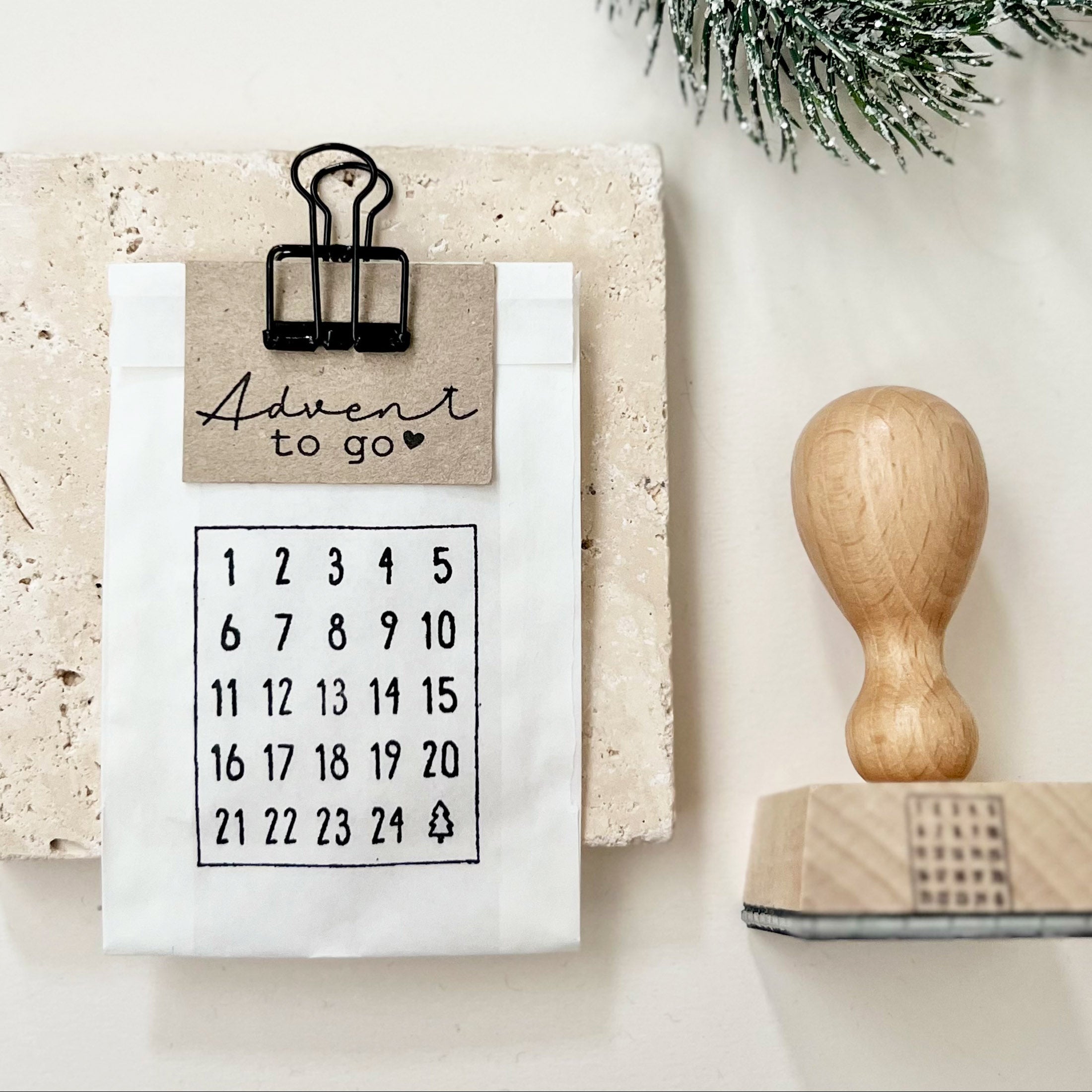Kleine weiße Pergamintüte, bestempelt mit den Zahlen 1 bis 24 und einem Mini-Tannenbaum. An das Tütchen ist ein kleiner Papieranhänger geklemmt, bestempelt mit dem Text "Advent to go". Rechts neben dem Tütchen liegt ein Holzstempel mit dem das Motiv "Adventskalenderzahlen" gestempelt wurde.
