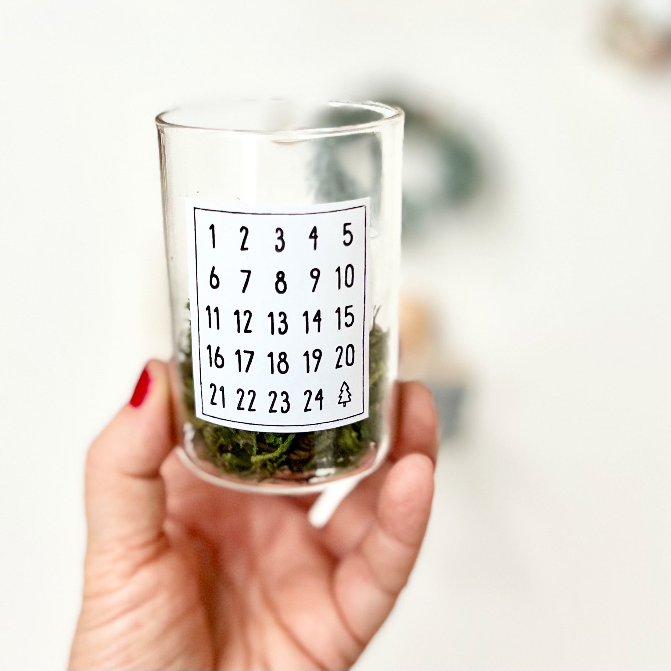 Kleines Glas, gefüllt mit Moos und einem Teelicht, in einer Hand gehalten. Auf dem Glas klebt ein Etikett, bestempelt mit den Zahlen 1 bis 24 und einem kleinen Tannenbaum. Den dafür verwendeten Weihnachtsstempel mit Motiv "Adventskalenderzahlen" sieht man auf vorherigem Foto.