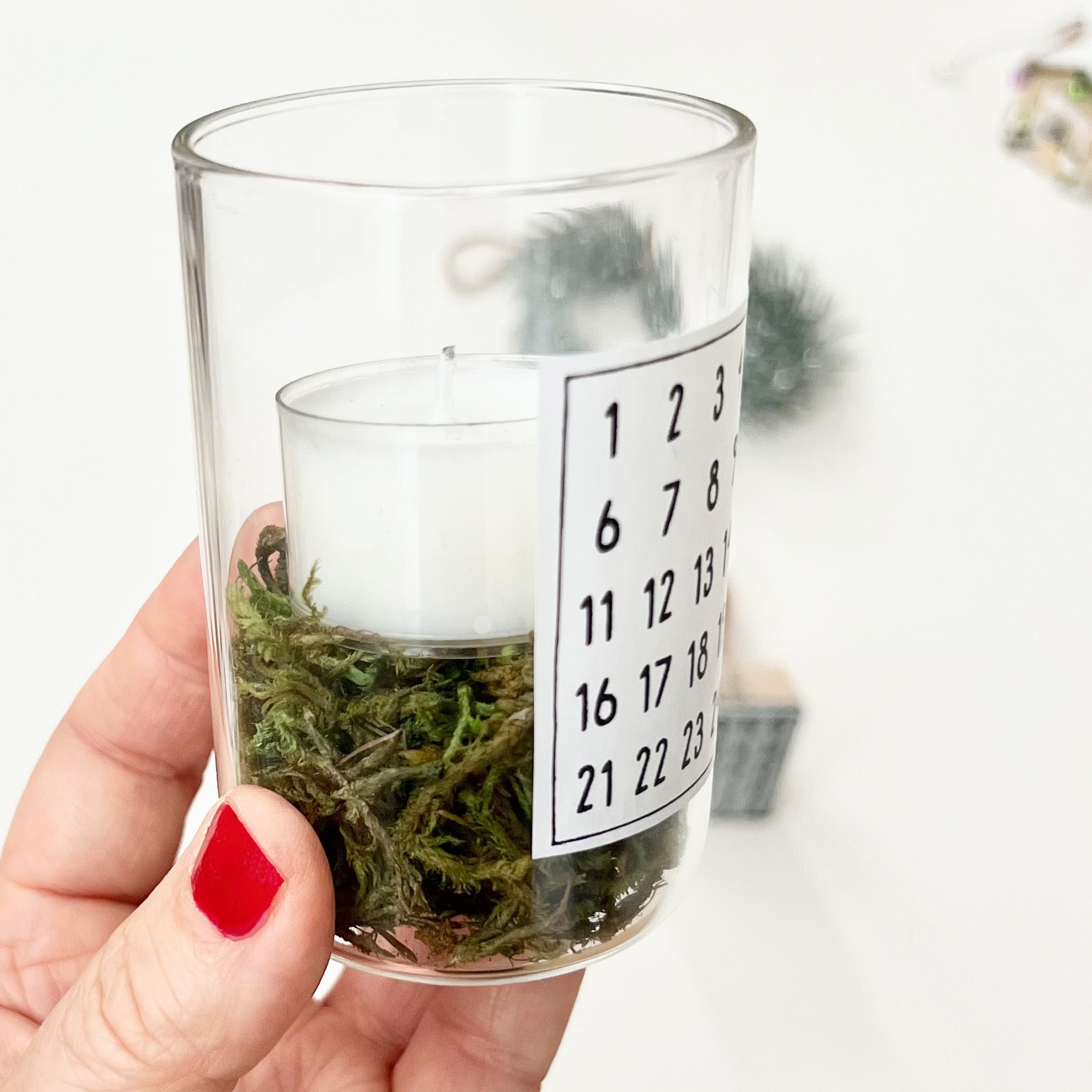 Kleines Glas, gefüllt mit Moos und einem Teelicht, in einer Hand gehalten. Auf dem Glas klebt ein Etikett, bestempelt mit den Zahlen 1 bis 24 und einem kleinen Tannenbaum. Den dafür verwendeten Weihnachtsstempel mit Motiv "Adventskalenderzahlen" sieht man auf dem ersten Artikelfoto.