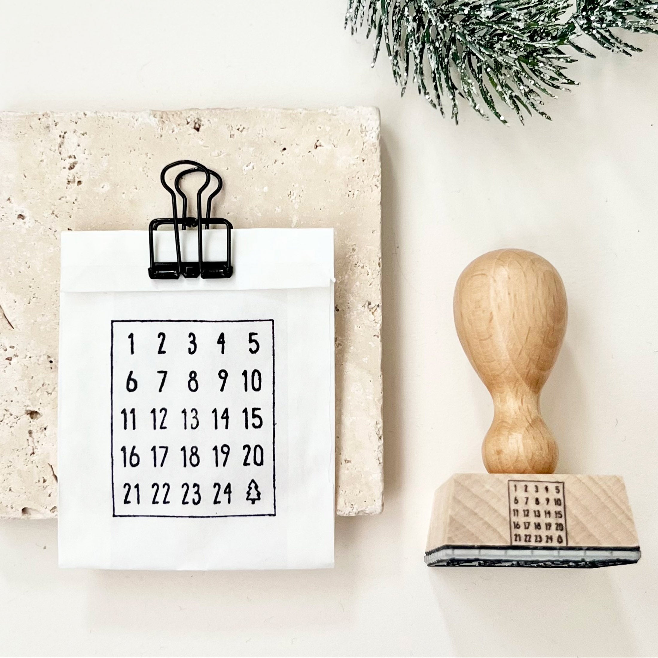 Weihnachtsstempel aus Holz mit Griff liegend, Motiv Adventskalenderzahlen, gestempelt auf eine kleine weiße Papiertüte, die verschlossen ist mit einer schwarzen Klammer.